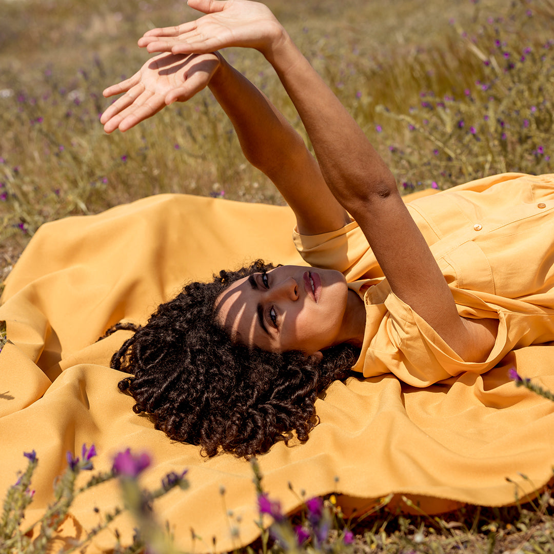 A person in a yellow outfit lies on a mustard blanket in a field of purple flowers, arms raised.