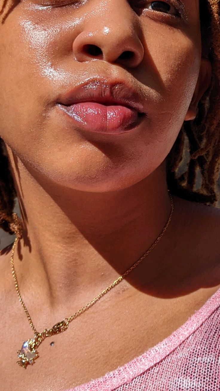 A person with dark skin applies a nude lipstick to their lips, with long dark hair visible.