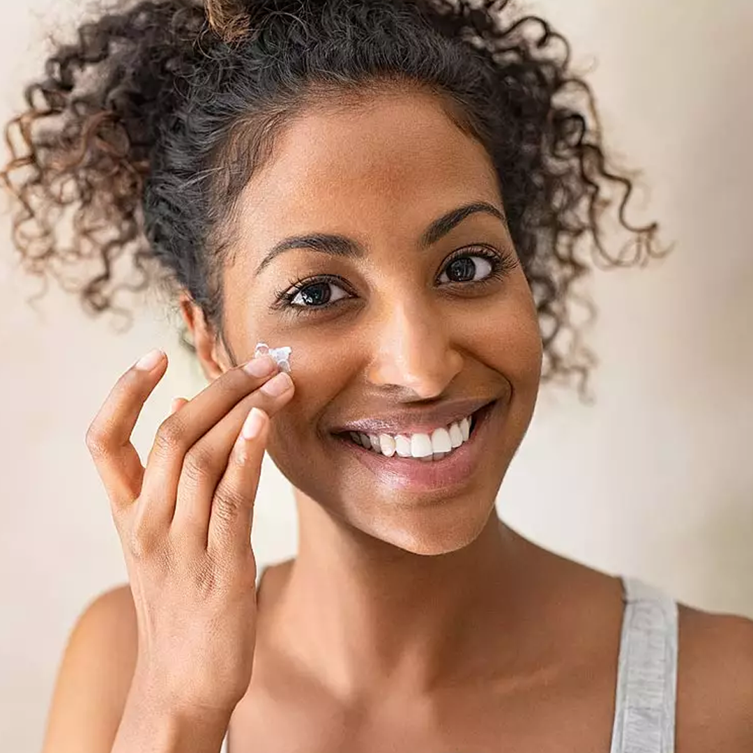 A person with curly hair applies cream to their cheek, smiling in a light gray top.