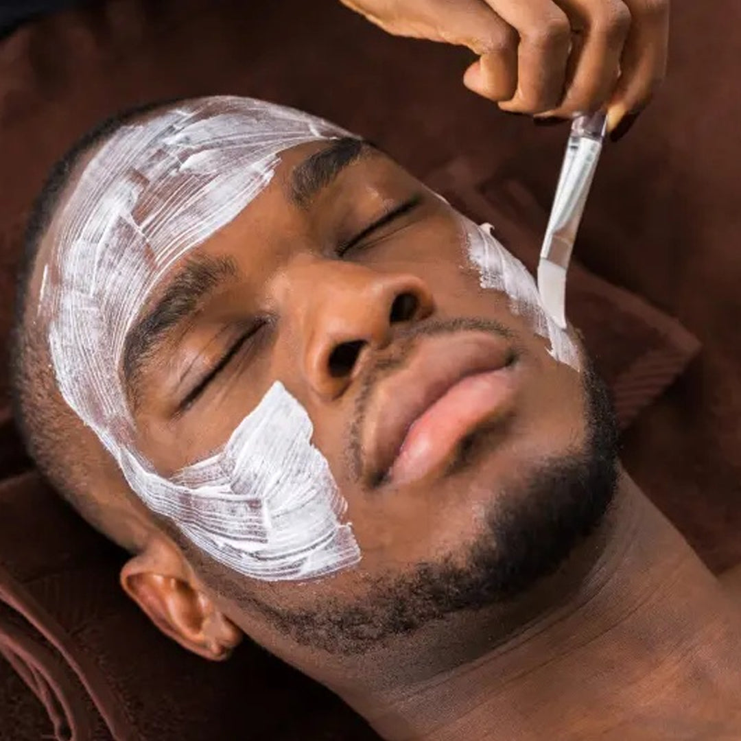 A person with a white face mask being applied by another using a spatula, on a brown surface.